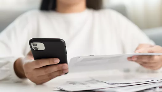 A person holding a black smartphone in one hand while reviewing a paper bill in the other.