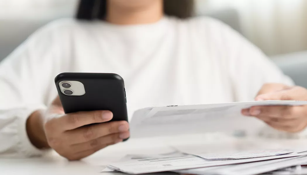 A person holding a black smartphone in one hand while reviewing a paper bill in the other.